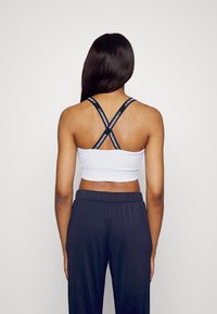 Bílý žebrovaný krátký top s černými překříženými ramínky nesoucími text s logem. Kombinován s tmavě modrými kalhotami, které mají hladkou texturu.