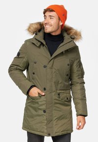 Chaqueta parka color caqui con capucha de piel sintética, cierre de botones y cremallera, múltiples bolsillos y puños de canalé. Gorro naranja visible.