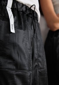 Black pants with a glossy finish, featuring an elastic waistband, drawstring, and distinct side pockets. The texture appears slightly wrinkled.