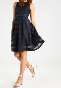 Mörkblå utanärmad klänning med en figurnära överdel och plisserad kjol. Har ett strukturerat mönster och transparenta horisontella ränder. Silverfärgade klacksandaler.