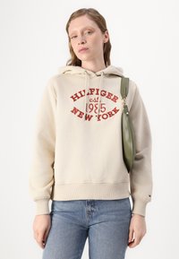 Felpa beige con cappuccio e stampa rossa "HILFIGER est. 1985 NEW YORK." Presenta polsini e orlo a coste. Abbinata a jeans blu e una borsa verde.