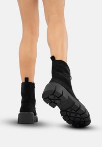 Botines de ante negro con suela de goma gruesa y suela texturizada, que presentan un cierre de cremallera lateral y una pestaña en la parte trasera.