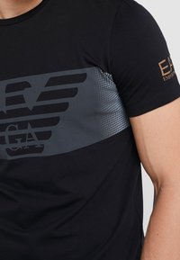 Musta t-paita, joka on valmistettu puuvillasta ja jossa on harmaa graafinen kuvio, jossa on viivoja ja pisteitä, sekä kultainen Emporio Armani -logo hihassa.