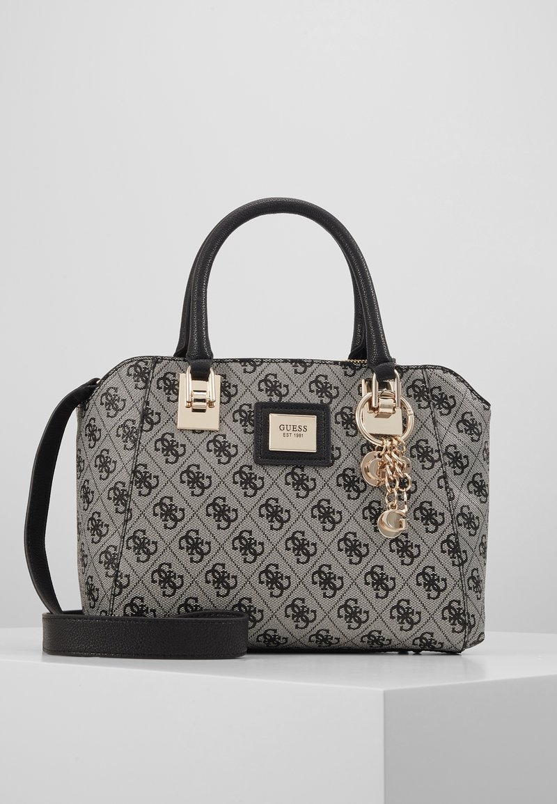 Sac à main Guess gris et noir à motifs avec poignées noires, ferrures dorées, breloques décoratives et bandoulière amovible sur une surface blanche.