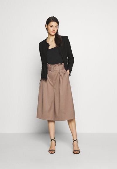 Veste noire texturée avec des bords effilochés, associée à un pantalon large beige taille haute et des sandales noires à lanières.