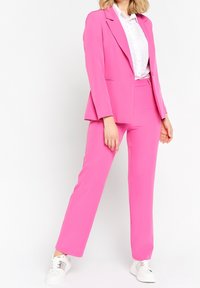 Bright pink suit with a tailored blazer and straight-leg trousers. Accompanied by a white button-up shirt with subtle texture and white sneakers.