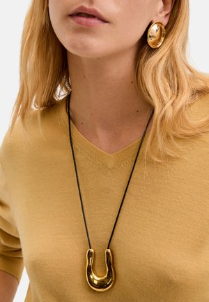 Pendentif en métal de couleur or en forme de U, suspendu à un cordon noir. Porté avec un pull en v jaune moutarde et des boucles d'oreilles circulaires assorties.