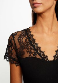 Bodysuit noir avec un décolleté et des manches en dentelle à motifs floraux, texture ribbed et bords festonnés.