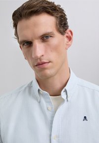 Lichtblauw gestreept overhemd met knopen en een geborduurd logo van een schedel met gekruiste beenderen. Voorzien van een klassieke kraag en knopen aan de mouwen.