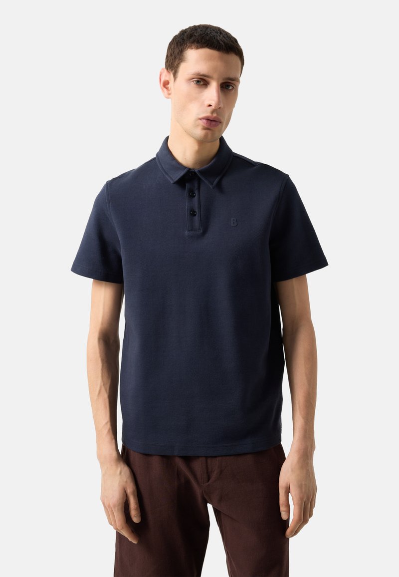 Jeune homme portant un polo bleu marine à manches courtes et un pantalon marron foncé, debout devant un fond clair uni.
