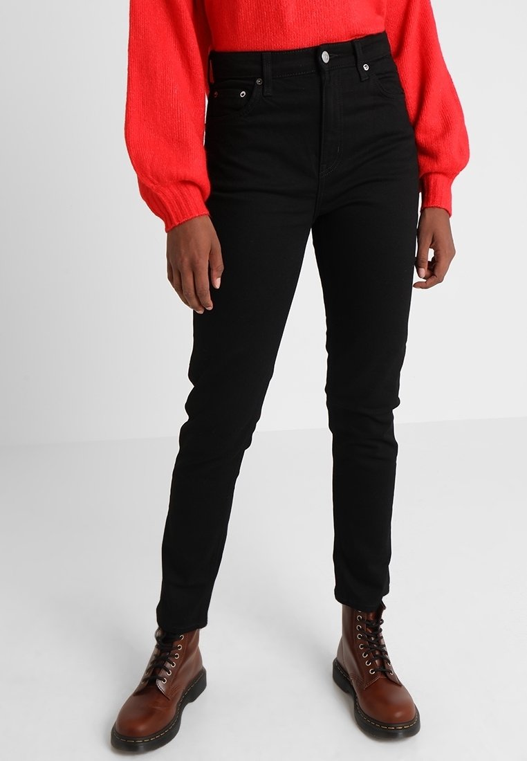 Personne portant un jean noir slim, des bottes marron à lacets et un pull à manches longues rouge vif sur un fond clair uni.