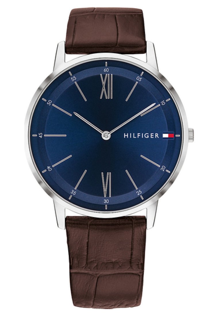 Reloj con correa de cuero marrón y caja de acero inoxidable plateado. La esfera redonda azul presenta marcadores de hora plateados, XII y VI en números romanos, y el logo de la marca.