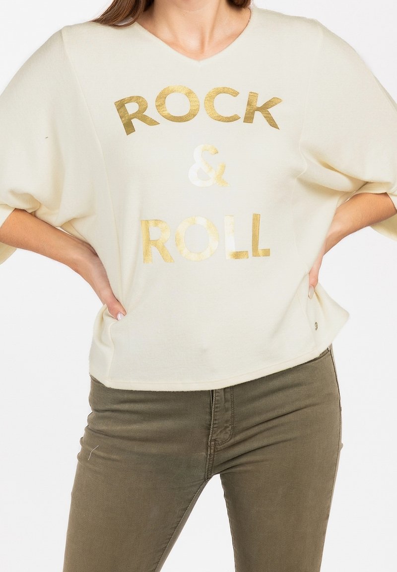 Femme portant un t-shirt crème à manches courtes avec le texte doré "ROCK & ROLL", associé à un pantalon ajusté vert olive, mains sur les hanches.