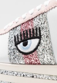 Vit sneaker med en tjock sula, med rosa, silver och glittriga detaljer. En broderad blå ögondesign med svarta fransar är framträdande.