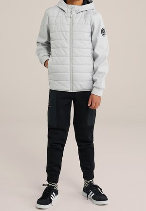 Bambino che indossa una giacca a vento con cappuccio grigio chiaro, pantaloni cargo neri, calzini a scacchi e sneakers nere, in piedi con le mani rilassate.