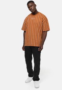 Brunstripet oversized t-skjorte med korte ermer og hvite vertikale linjer, kombinert med svarte slitte cargobukser og hvite sneakers.