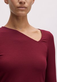 Burgunderfarbene, gerippte Langarmbluse mit asymmetrischem Halsausschnitt und taillierter Silhouette, aus weichem, strukturiertem Stoff gefertigt.