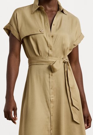 Dunkelhäutige Person, die ein beiges Hemdkleid mit kurzen hochgekrempelten Ärmeln und einem gebundenen Taillengürtel trägt.