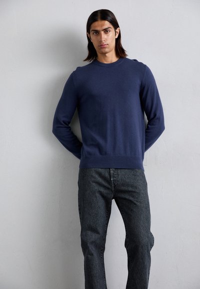 Young man with long dark hair wearing a navy crewneck sweater and dark gray jeans, standing against a plain off-white wall.