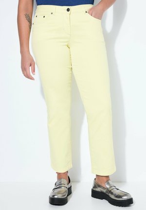 Personne portant un pantalon jaune pâle avec poches avant, un tatouage visible sur le bras gauche, un haut bleu foncé, et des mocassins à plateforme argentés.