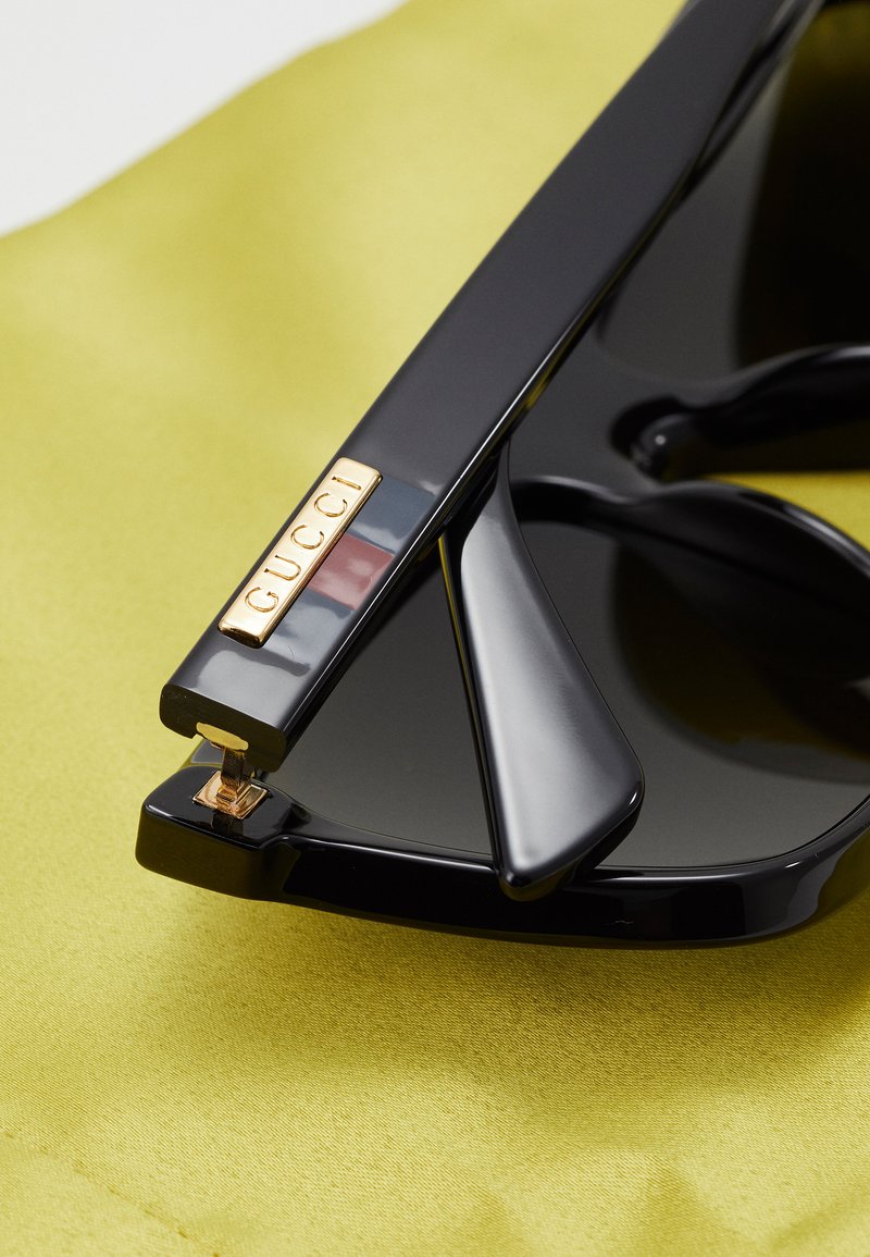 Sorte solbriller med en blank finish og signatur guld hardware detalje med mærkets navn. Glassene er mørke og har en vinkelret form.