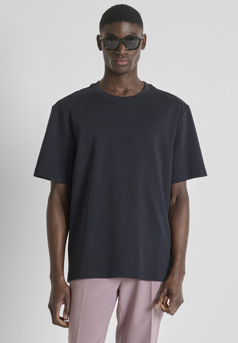 Homme portant un T-shirt noir surdimensionné et un pantalon lilas avec des lunettes de soleil noires, debout devant un fond gris clair uni.