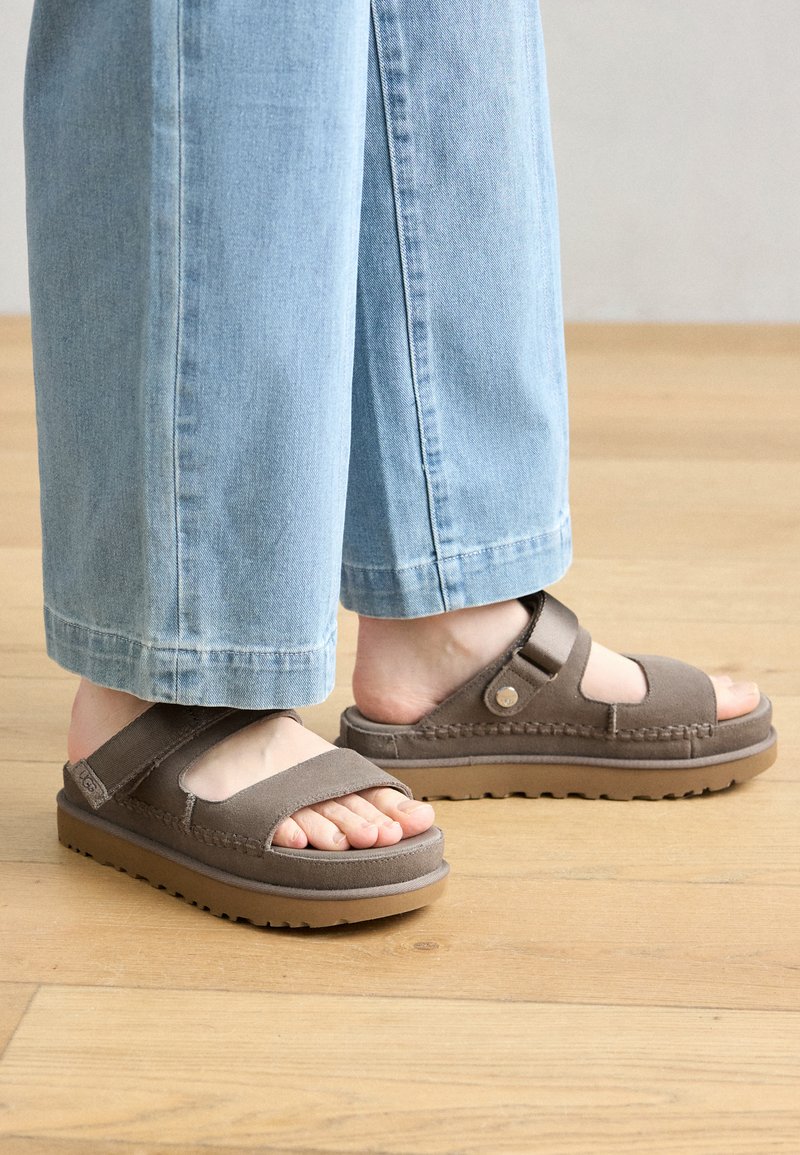 Brązowe zamszowe sandały z szerokimi paskami, regulowanym zapięciem i grubą gumową podeszwą, połączone z jasnoniebieskimi spodniami dżinsowymi. Wnętrze.