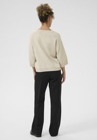 Beige gebreide trui met driekwart mouwen, geribbelde halslijn en golvende zoom. Gekleed met zwarte broeken. Gezien van achteren.