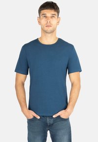Blaues Baumwoll-T-Shirt mit rundem Ausschnitt, kurzen Ärmeln und einem dezenten Logo auf der Brust. Kombiniert mit ausgewaschenen blauen Jeans.