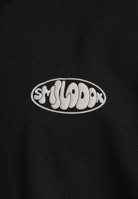Tejido de algodón negro con un logo ovalado blanco que contiene la palabra "SMILADOK" en una fuente redondeada y juguetona.
