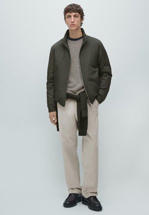 Giovane uomo con giacca oliva con zip sopra maglione grigio e maglione nero annodato in vita, pantaloni beige e scarpe nere, in piedi davanti a uno sfondo neutro.