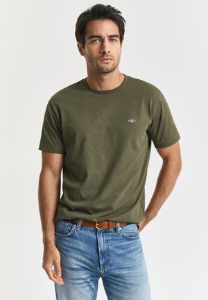 Mann iført olivengrønn kortermet t-skjorte og lyseblå jeans med brun belte, stående foran en ensfarget hvit bakgrunn.