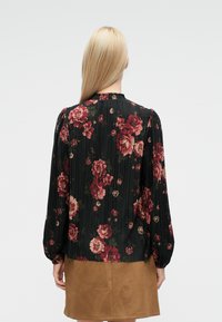 Svart blomstrete bluse med folder, med røde og beige blomster, lange ermer og en volangkant på halsen. Kombinert med et tan-farget skjørt.