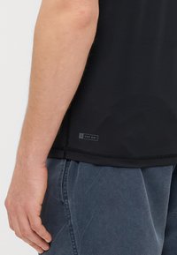 Svart UPF 50+ skjorta med korta ärmar, mjuk textur och platta sömmar, i kombination med grå byxor i en avslappnad passform.