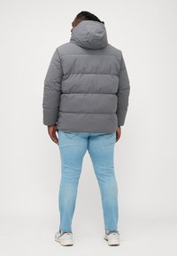 Doudoune grise avec un col montant et une capuche, présentant un motif matelassé. Associée à un jean bleu clair et des baskets grises.