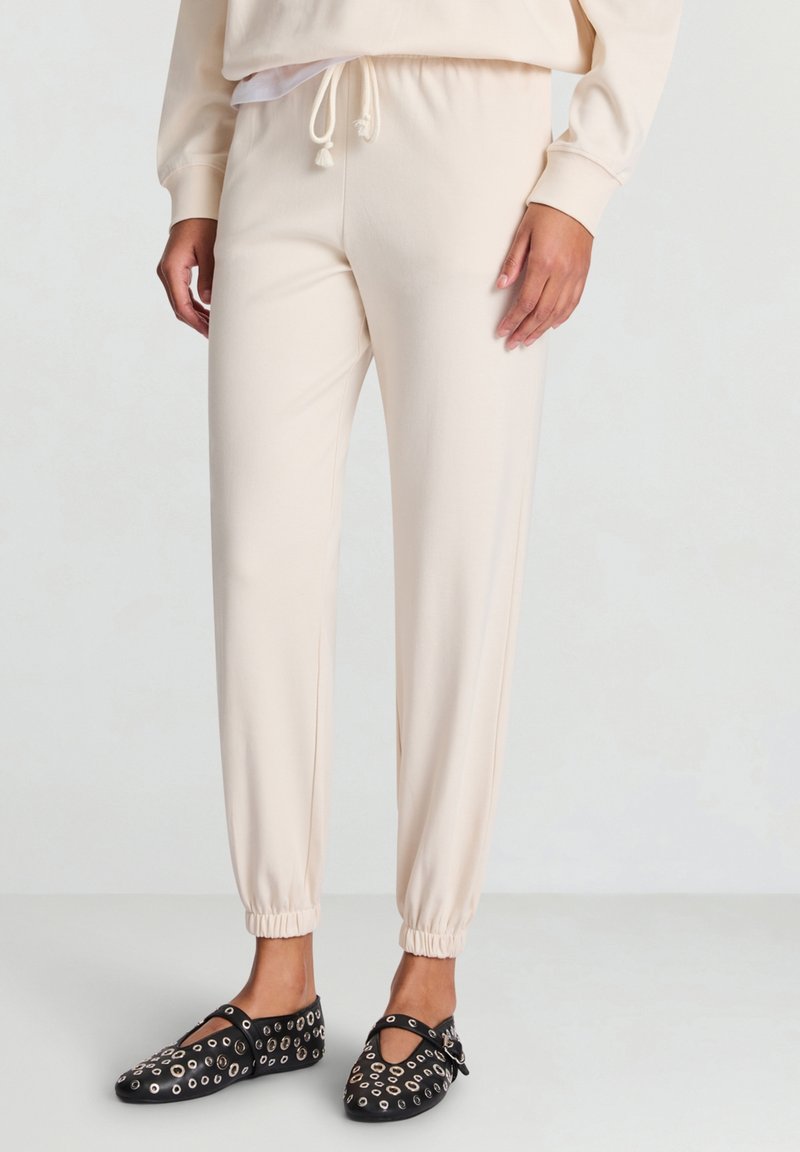 Pantaloni della tuta color crema con polsini elastici, vita con coulisse e consistenza liscia, abbinati a ballerine nere con occhielli argentati.