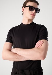 Fekete, rövid ujjú póló, magas gallérral, sima anyagból. Nagy fekete napszemüvegben viselve, karok keresztbe téve, semleges háttérrel.