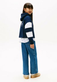 Granatowy skrócony hoodie z białymi paskami na rękawach, noszony na białej koszuli w kropki, zestawiony z szerokimi niebieskimi dżinsami i beżowymi sneakerami.