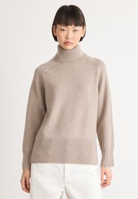 Giovane donna con capelli corti e scuri che indossa un maglione a collo alto beige e pantaloni bianchi su uno sfondo chiaro e semplice.