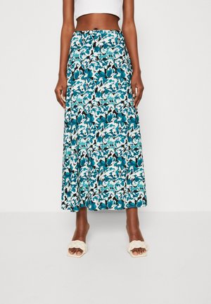 Femme portant une jupe taille haute, longueur cheville, avec un motif floral bleu, noir et blanc, et des sandales blanches tressées à enfiler.