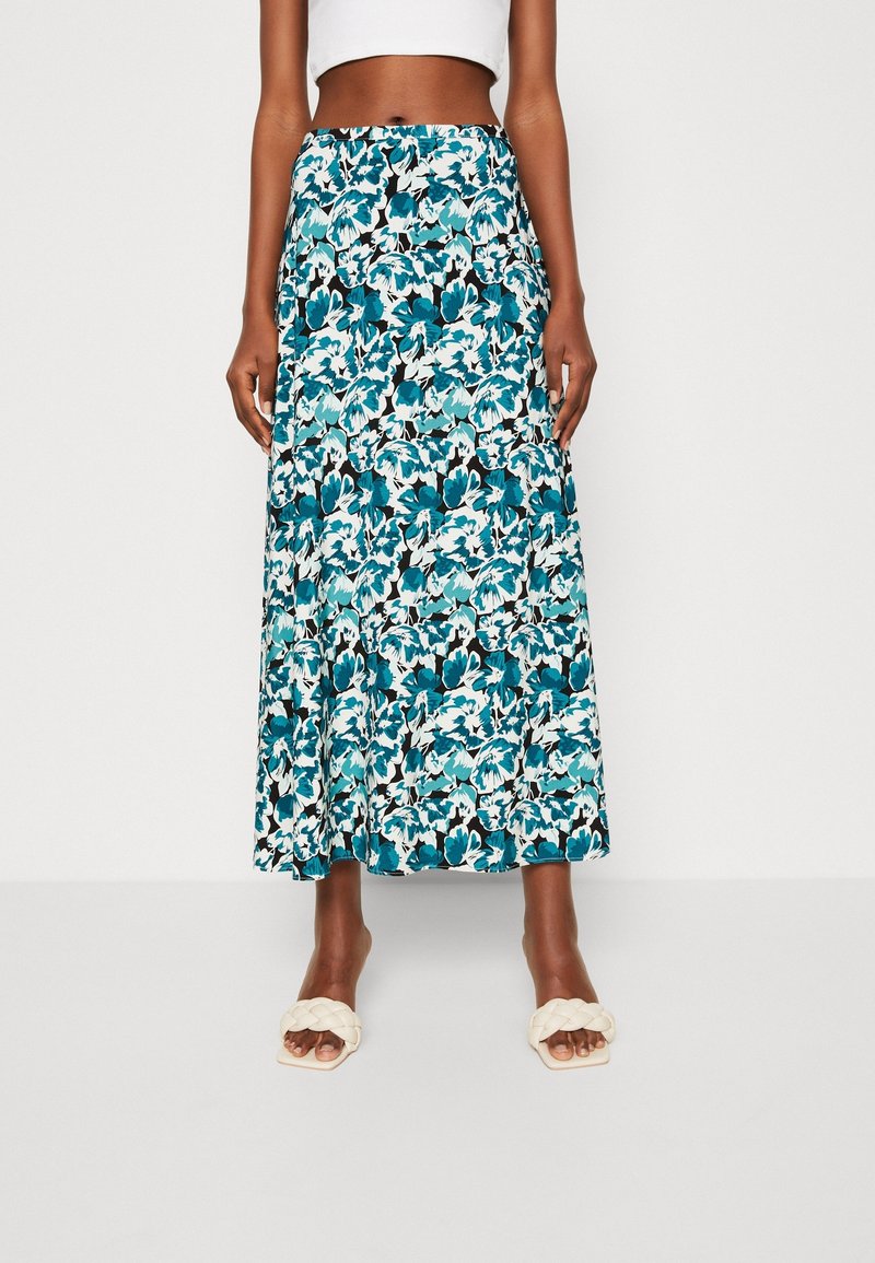 Femme portant une jupe taille haute, longueur cheville, avec un motif floral bleu, noir et blanc, et des sandales blanches tressées à enfiler.