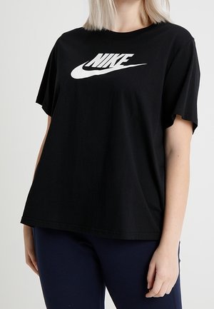 Person trägt ein lockeres schwarzes Nike T-Shirt mit weißem Logo, kombiniert mit dunkelblauer Hose, stehend vor einem schlichten Hintergrund.