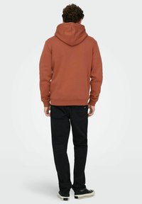 Man draagt een roestkleurige hoodie, zwarte spijkerbroek en zwarte sneakers, staat met de rug naar de kijker op een effen witte achtergrond.