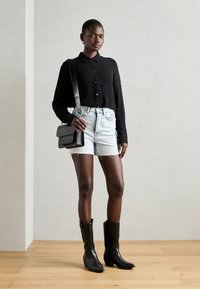 Maglietta nera a maniche lunghe, pantaloni shorts in denim con orli sfrangiati, borsa a tracolla nera con finitura testurizzata e stivali cowboy neri con punta affusolata.