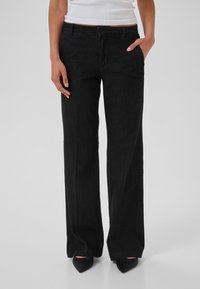 Mustat, leveät denim-housut, joissa on sileä rakenne, klassinen viiden taskun muotoilu ja vetoketjusuljenta, yhdistettynä teräväkärkisiin mustiin kenkiin.