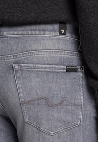 Jeans in denim grigio con una tasca posteriore che presenta un motivo di cucitura curvo. Un'etichetta nera vicino alla vita recita "per tutta l'umanità".