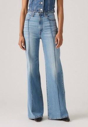 Donna che indossa jeans svasati a vita alta di un blu chiaro, con cuciture verticali frontali, e scarpe nere a punta su uno sfondo semplice.