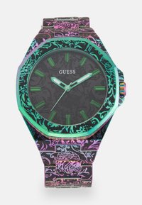 Montre noire octogonale avec un bracelet à motif, affichant des index et des aiguilles verts, ainsi qu'une lunette irisée. Nom de la marque "Guess" affiché.
