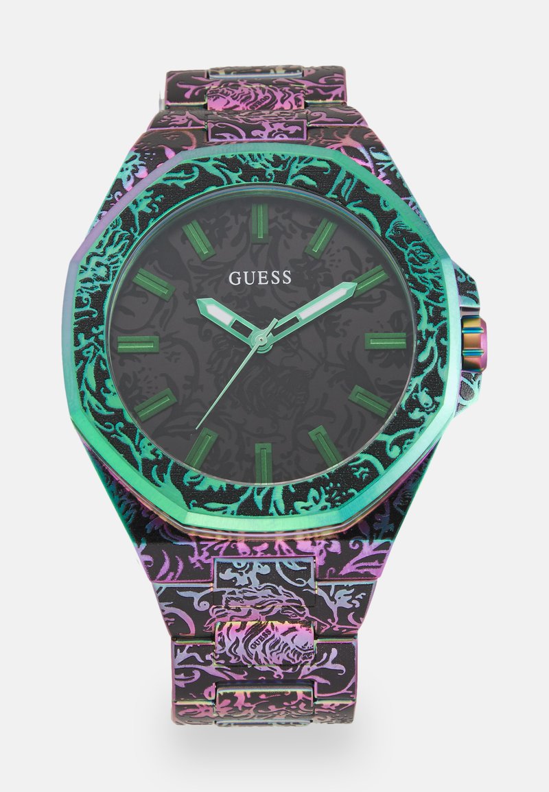 Montre noire octogonale avec un bracelet à motif, affichant des index et des aiguilles verts, ainsi qu'une lunette irisée. Nom de la marque "Guess" affiché.