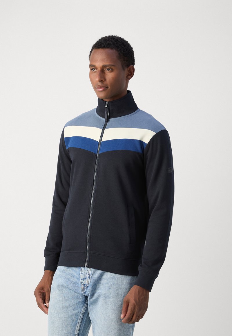 Sweatshirt zippé marine avec col montant, présentant des rayures horizontales en bleu clair, blanc et bleu foncé. Tissu en mélange de coton lisse.